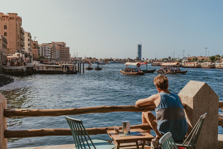 dubai creek, waterfront, man sitting, abra boats, traditional boats, dubai water taxis, relaxing view, outdoor seating, creek view, people, abra ride, sunny day, sightseeing, waterfront cafe, boat ride, casual scene, nature, cityscape, urban view, waterfront dining, riverfront, dubai, pier, sea, outdoors, boat